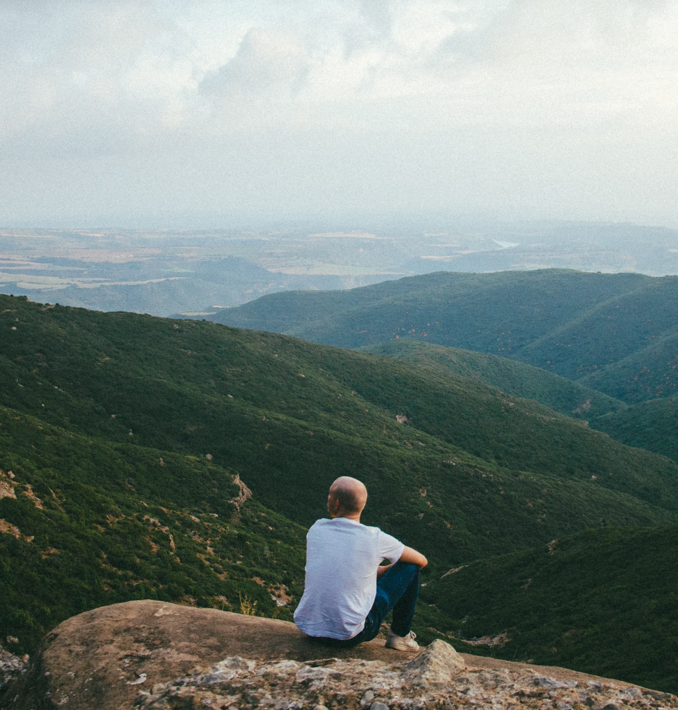 man sitting in field, looking to mountains